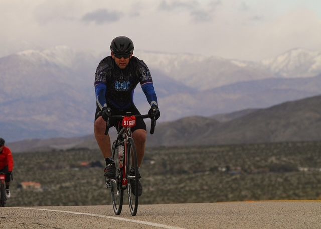 Aaron riding a road bike with a race number pinned on during a desert century ride, snow-capped mountains in the distance