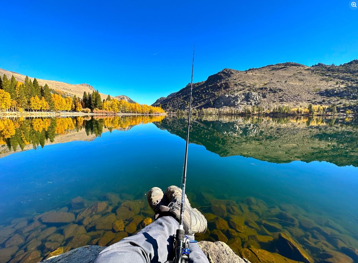 Aaron's boots propped on a rock beside a glassy alpine lake in autumn, fishing rod resting against his leg, golden aspens and granite reflected in the still water
