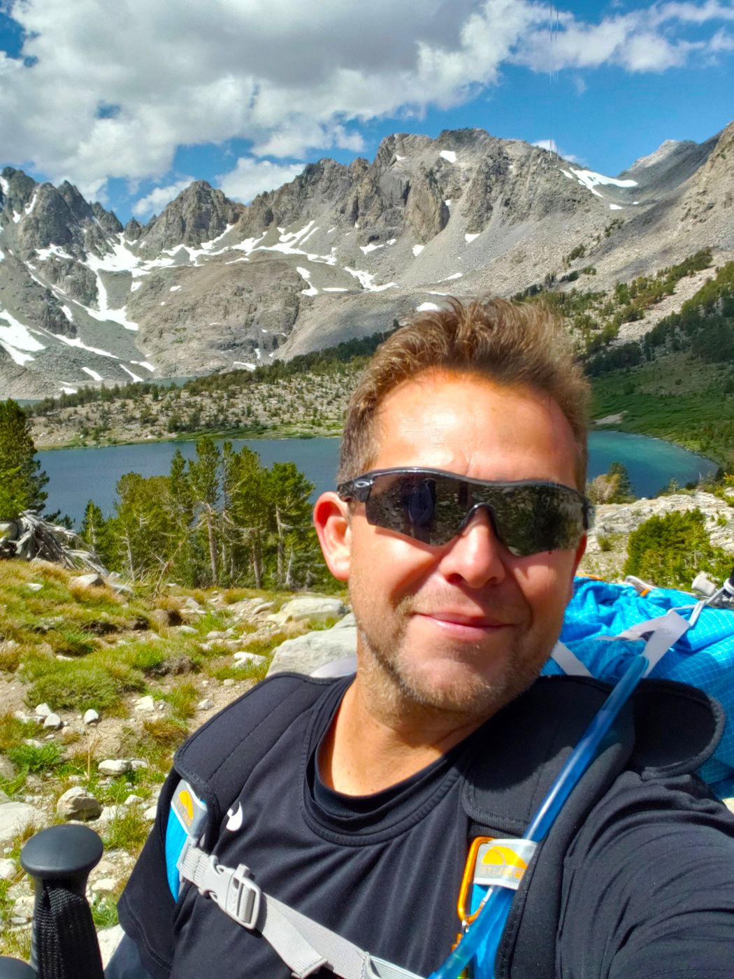 Aaron on a High Sierra trail above an alpine lake, granite peaks with patches of snow behind him