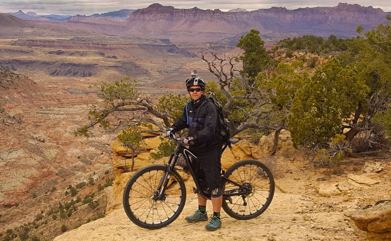 Aaron with his mountain bike on a red rock canyon rim, desert cliffs and junipers behind him