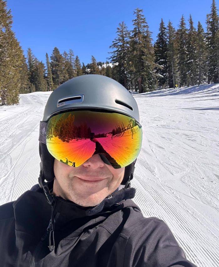 Aaron in helmet and mirrored goggles on a freshly groomed run, pine trees and blue sky behind